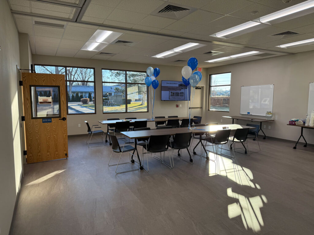 Large room with several windows. Two long rectangular tables with 10 chairs around each table fill the room.